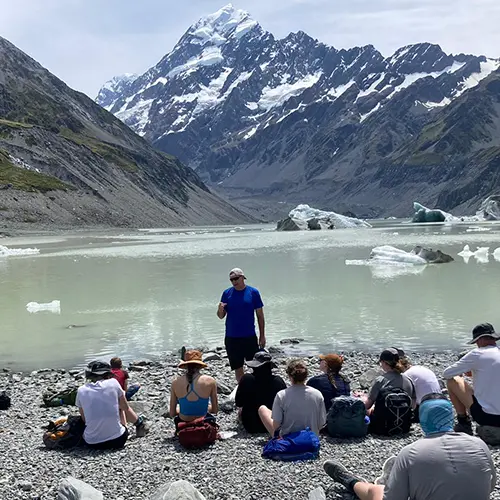 Professor Cottrell talking to a group of students at an outdoor program overlooking a lake and mountain range.