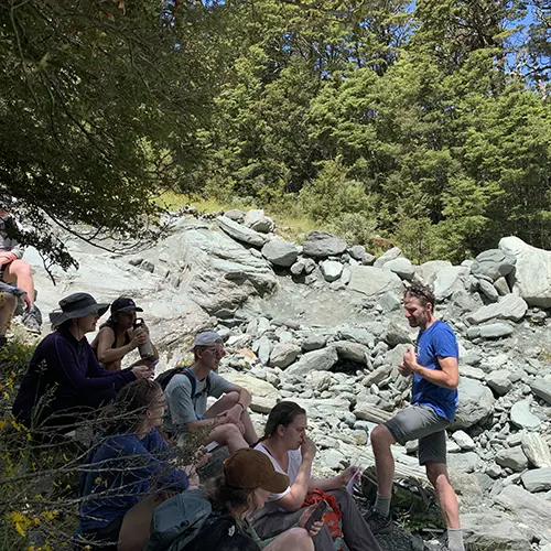 Syring talking to a group of student at an outdoor program, surrounded by rocks and trees.