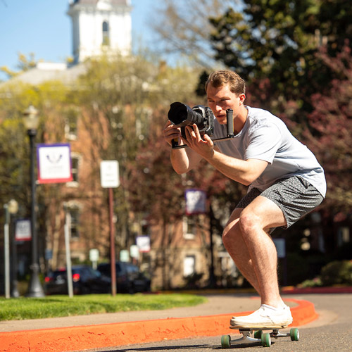 a student filming video while riding a skateboard on campus