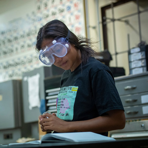 female student in the chemistry lab with goggles on reading experiment instructions from a textbook