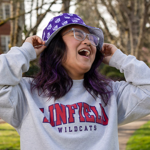 Female student wearing Linfield hoodie and hat, laughing.