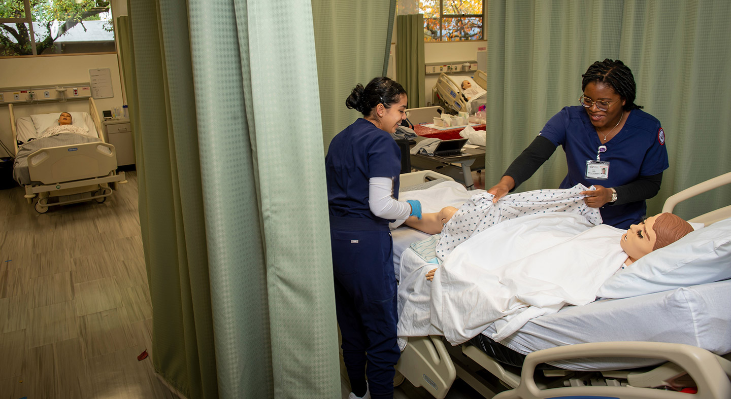 Two nursing students in the simulation lab practicing with a manikin lying in a hospital bed.