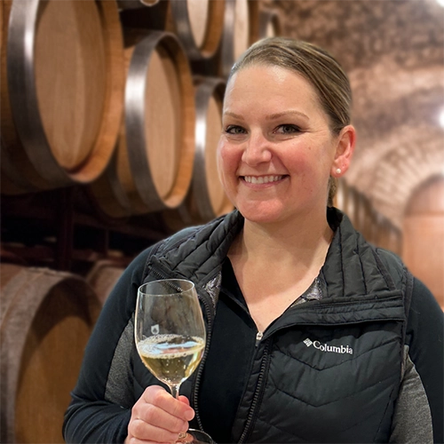 student holding a glass of wine in front of wine barrels.