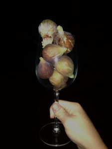 A hand holds a clear wine glass filled with figs of different colors against a black backdrop