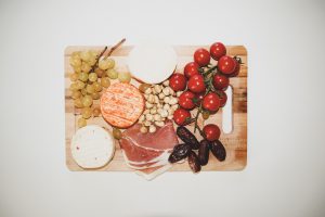 a wooden tray filled with grapes, prosciutto, cheeses, figs, and cherry tomatoes against a white backdrop