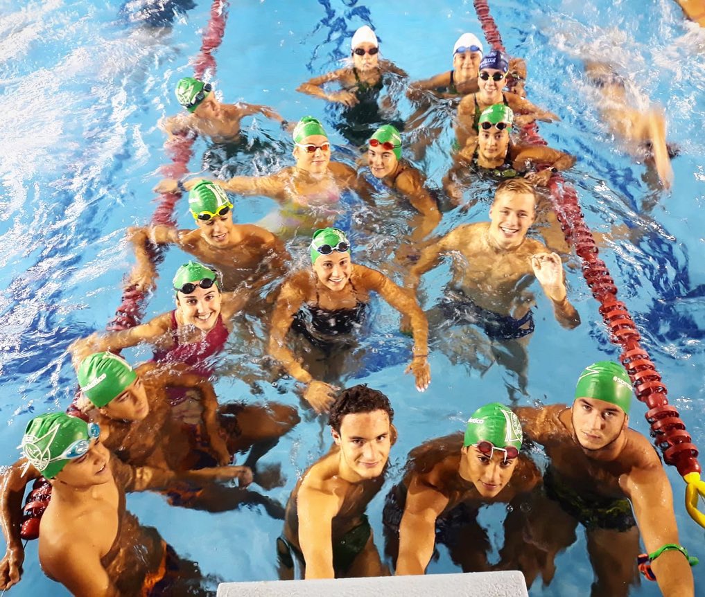 A group of college-aged men and women treading water in the swim lane of a pool. A majority of them are in green swim caps with goggles.