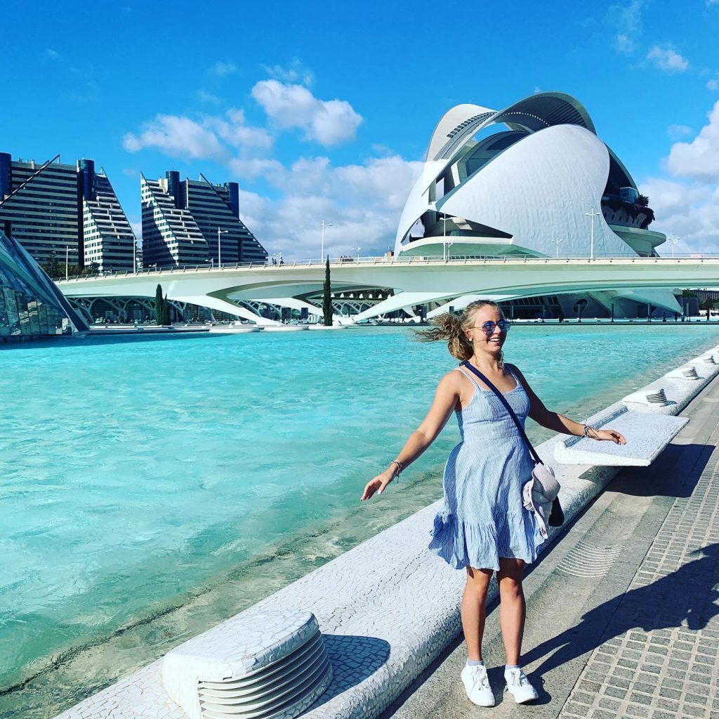 A girl in a blue dress stands in front of a light teal body of water. There is an urban white bridge and building in the distance under a blue sky.