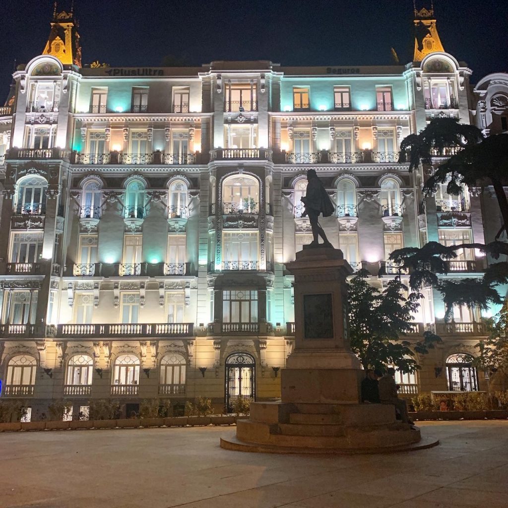 A six story building in Madrid, Spain against a clear night sky. The lights on the building are a mix of warm yellow lights and teal lights shining on the front of the building. There is a stature of a man in front of the building.