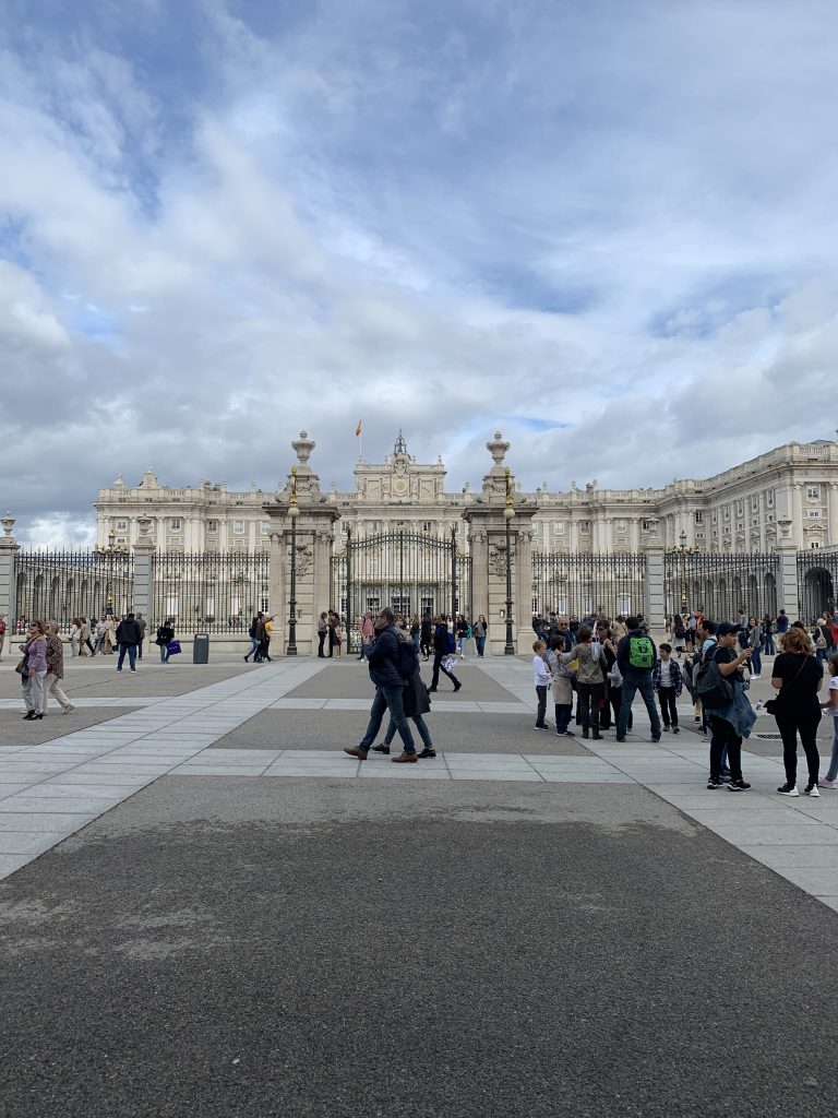 People walk past a large white European palace surrounded by a fence with a grand gate.