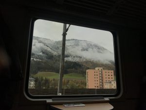 View from the train to Obertraun of tall buildings with cloud covered mountains in the background.