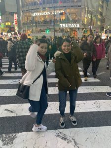 Two girls standing in Shibuya Crossing with people crossing the street behind them.