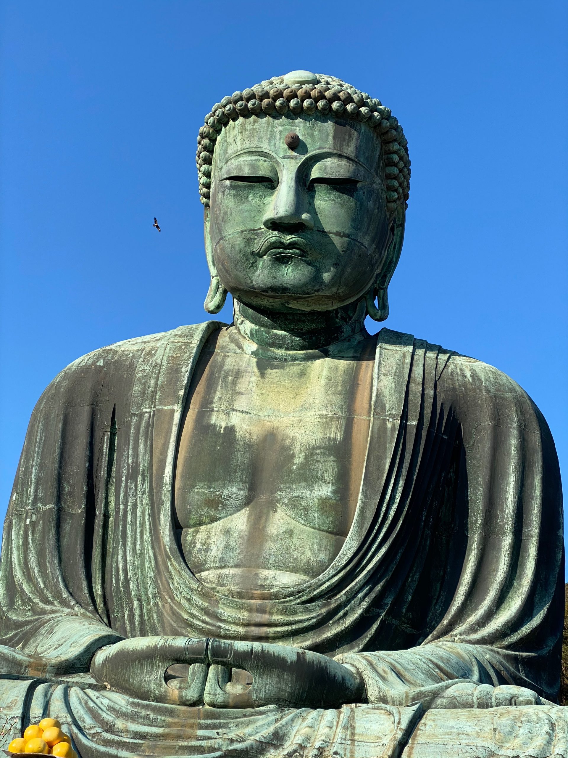 A gigantic statue of buddha with a blue sky behind it and some fruit by its legs