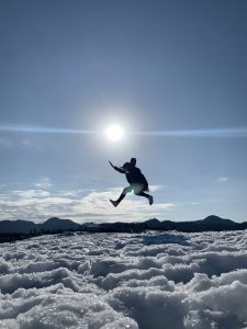 Girl jumping above snow, blue sunny sky behind her.