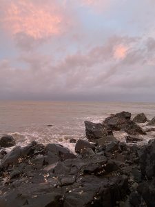 The ocean coming up to dark colored rocks with a sunrise in the sky behind them. The sky is pink and blue with a few clouds.