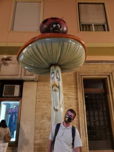 The author, pictured in a beige stiped polo, doing a silly pose beneath a mushroom statue, which has a ladybug statue on top. 