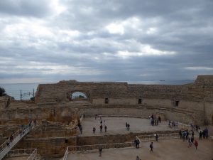 A view of the Roman amphitheater of Tarragona. A handful of tourists walk on the amphitheater. Gray clouds cover the sky. The ocean is in the background. 