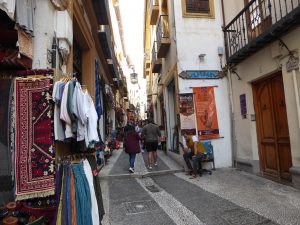 A street in the Arabic shopping district of Granada. Traditional dresses and rugs are in the foreground. People are walking on the cobblestone street with tall buildings looming over them.