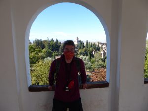 The author wearing a maroon jacket in front of an arched lookout point at La Alhambra. In the background is vegetation and a palace.