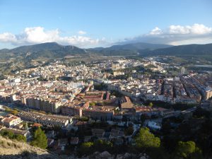 A panoramic view of the city of Alcoi from above. Many light-colored buildings compose the landscape. Some mountains are in the background, along with some white clouds. 