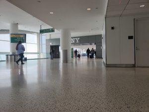 JFK NYC airport. The floor is white and reflecting a dim sunlight. Some passengers are seen walking around. A sign says NY.