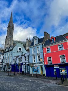Colorful 3-story row houses line the streets of Cobh with a tall church spire in the background.